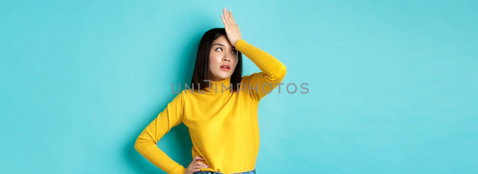 Emotions and lifestyle concept. Annoyed asian girl roll eyes and face palm, standing bothered in yellow pullover against blue background.