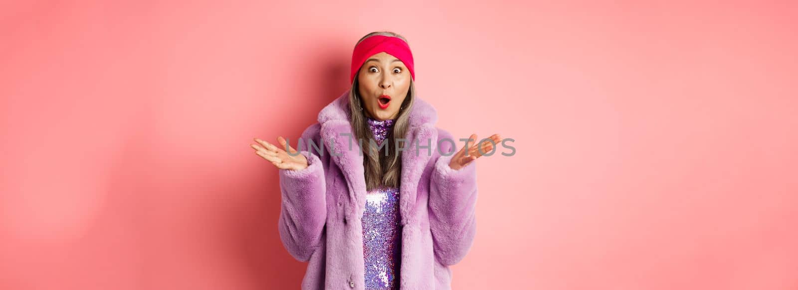 Fashion and shopping concept. Surprised elderly asian woman in stylish fake-fur coat and headband looking amazed at camera, rejoicing of good news, pink background.
