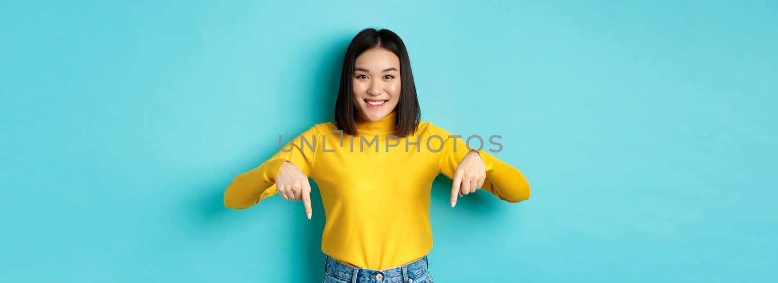 Shopping concept. Beautiful korean girl with happy smile, pointing fingers down at banner, standing against blue background.