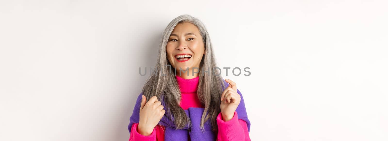 Close-up of fashionable asian senior woman, laughing and having fun, looking left cheerful, standing over white background.