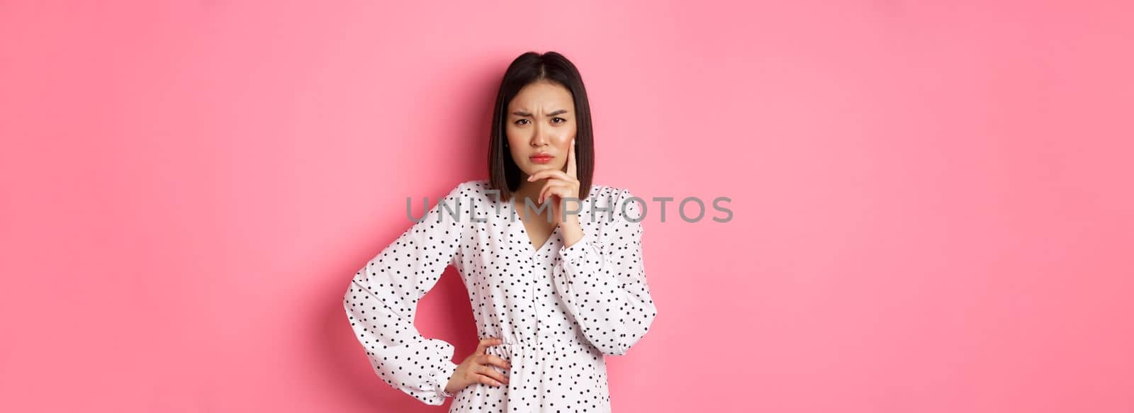 Concerned asian woman staring at camera suspicious, having doubts, thinking about something with serious face, standing against pink background.