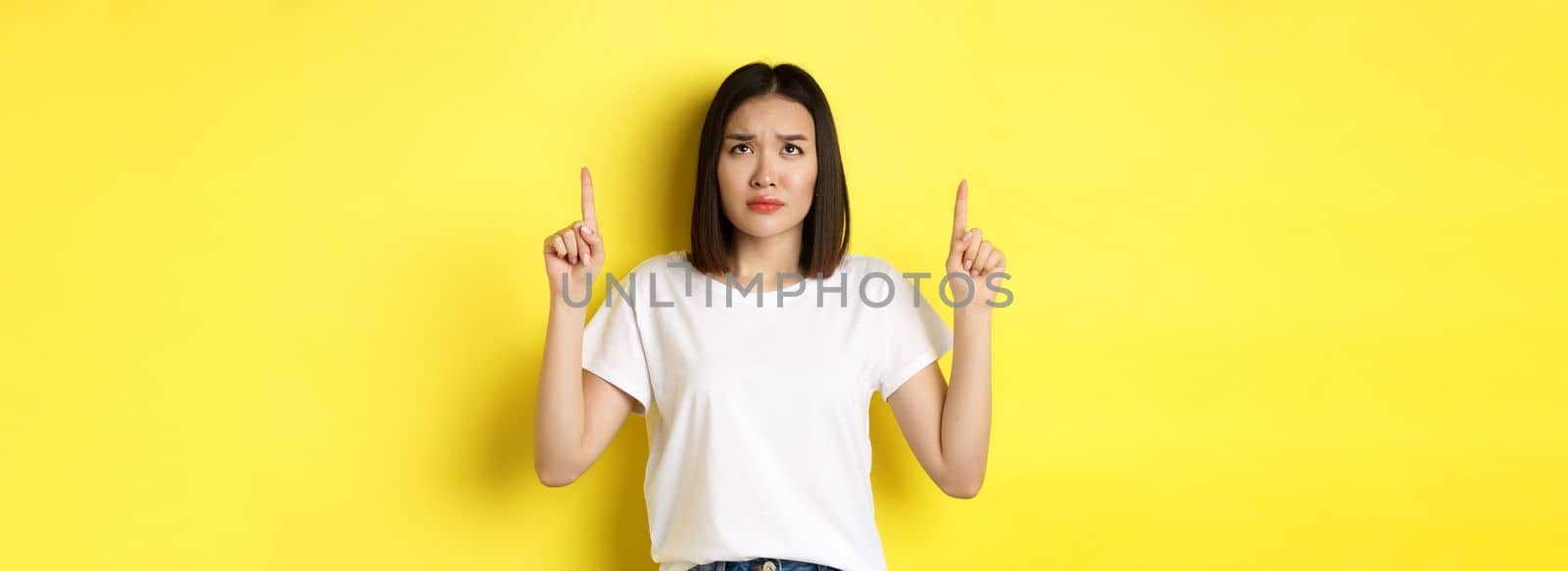 Beauty and fashion concept. Beautiful asian woman in white t-shirt pointing fingers up, standing over yellow background.