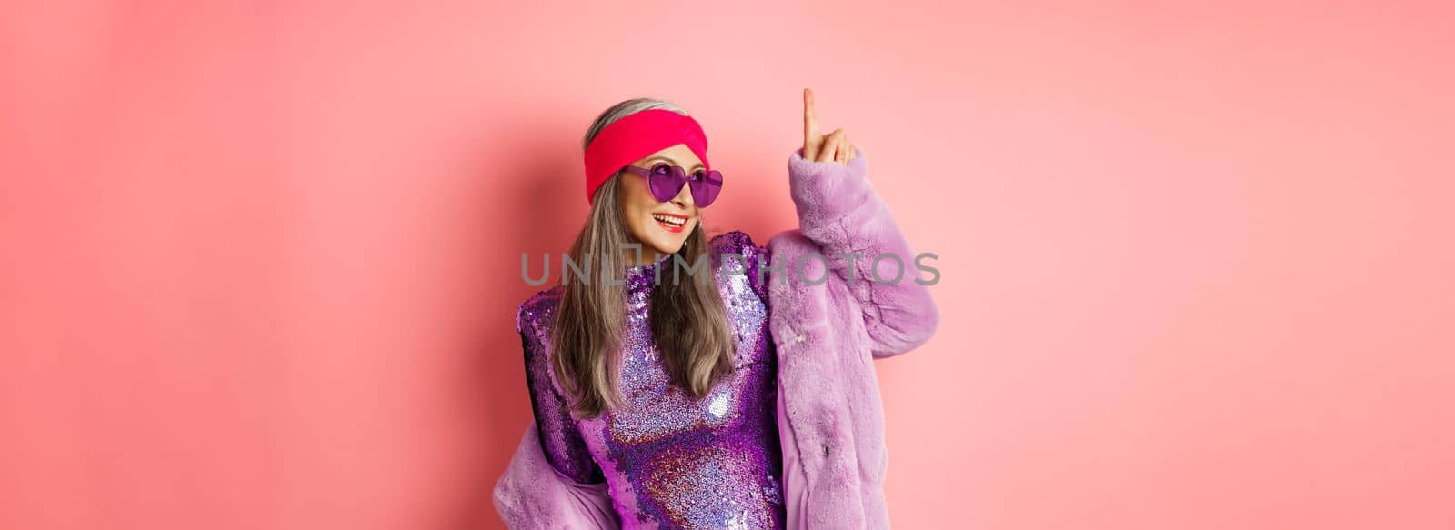 Asian old woman in fashionable glittering dress and purple faux fur coat dancing disco, pointing finger up and having fun, pink background.