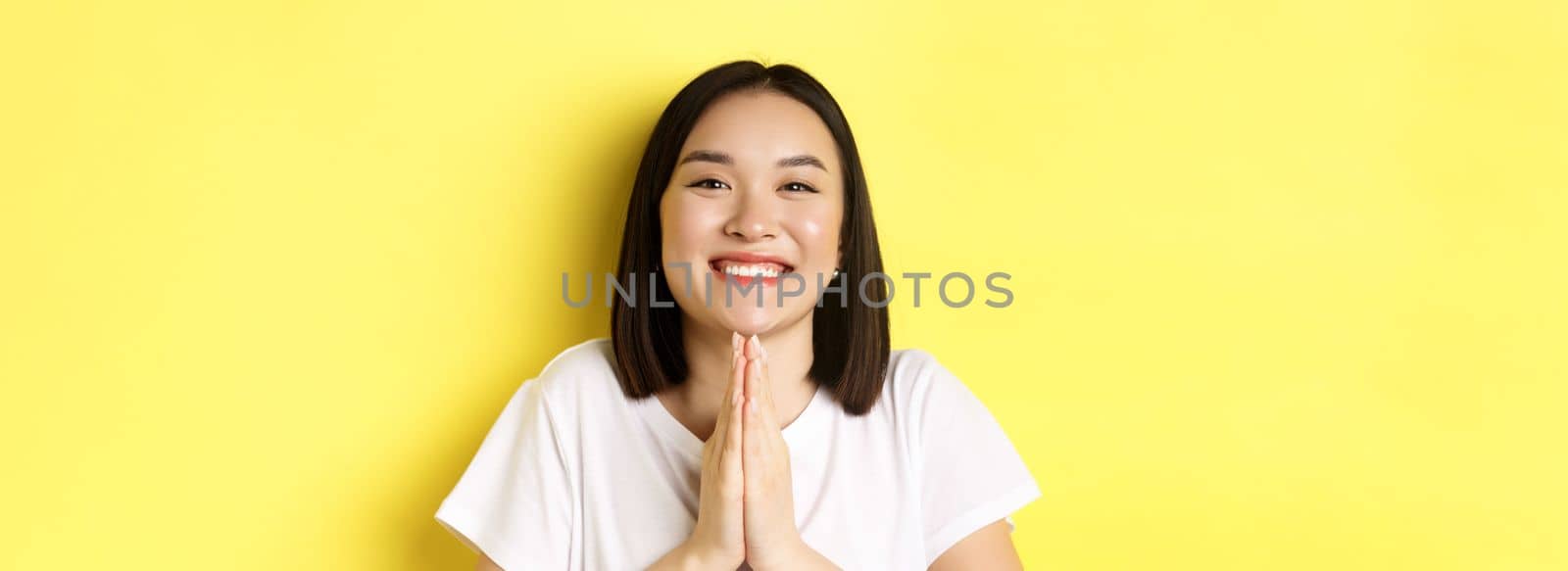 Close up of cute asian woman saying thank you and smiling, holding hands in namaste, pray gesture, standing over yellow background.