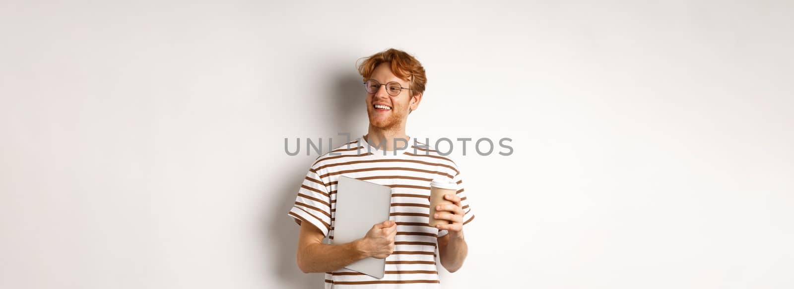 Handsome redhead male employee in glasses having break, drinking coffee and holding laptop, standing over white background.