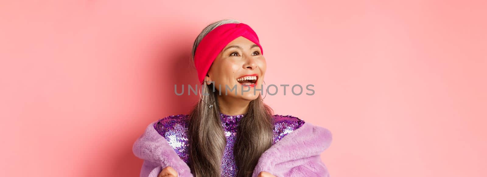 Fashion. Close-up of happy asian senior woman smiling, looking left with cheerful face, standing in purple faux fur coat, pink background.