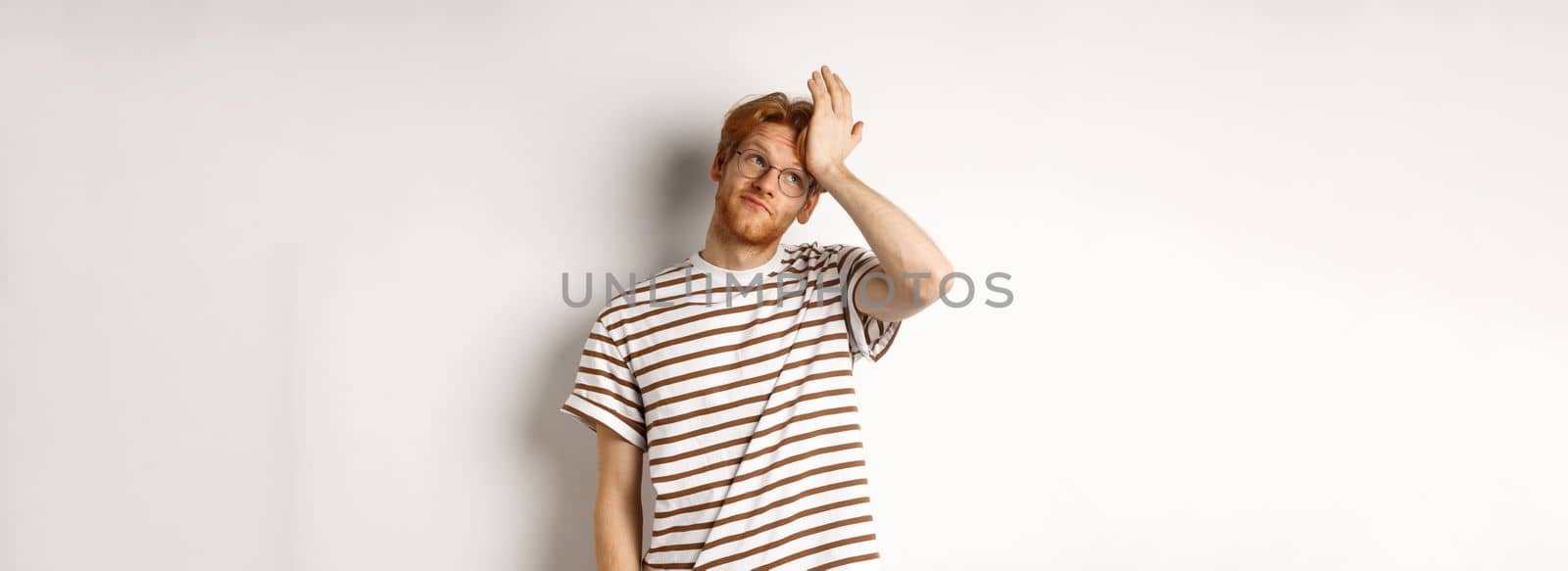 Tired young man with red hair and glasses, roll eyes and making facepalm bothered, standing over white background.