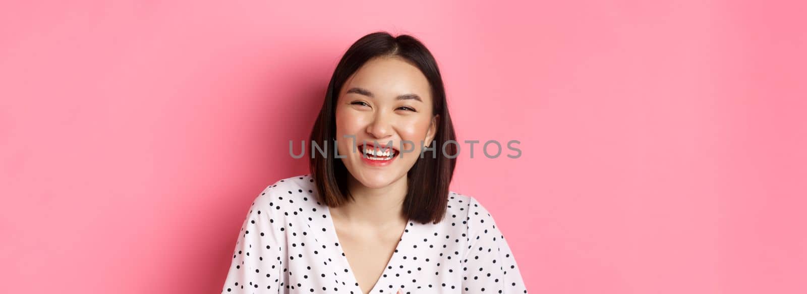 Beauty and lifestyle concept. Close-up of happy asian woman laughing and having fun, standing over pink background. Copy space