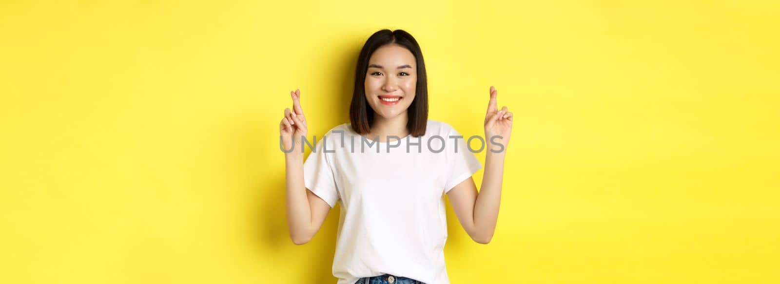 Hopeful asian girl making wish, cross fingers for good luck and praying with eyes closed, standing over yellow background.