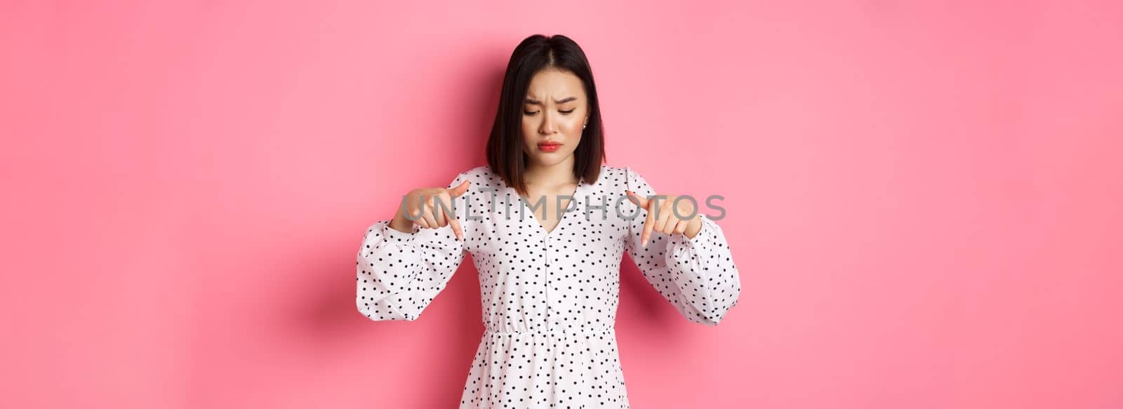 Concerned asian woman peeking down and pointing hand at disturbing banner, frowning upset, standing over pink background by Benzoix