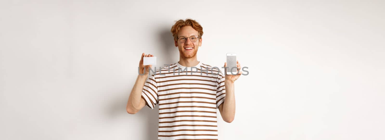 Shopping and finance concept. Young man showing blank mobile screen and plastic credit card, smiling at camera, white background.