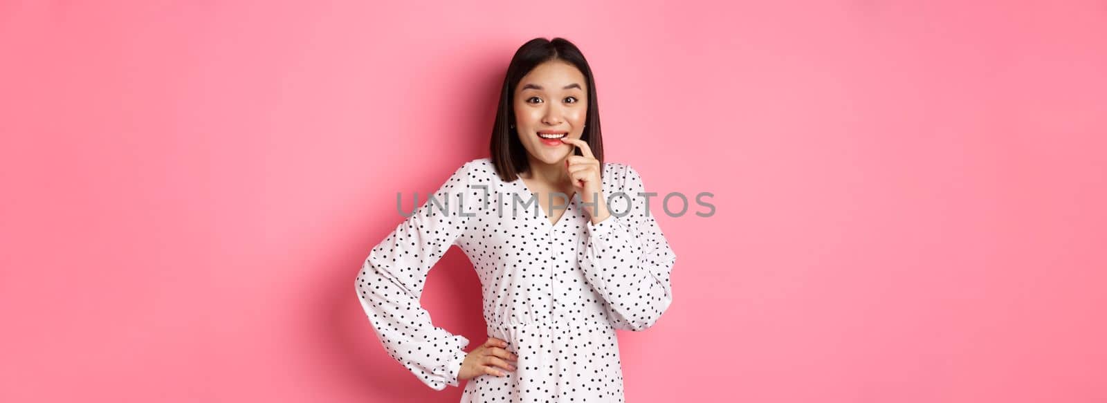 Image of beautiful young lady in dress, biting finger and smiling, staring with temptation and amazement, want buy something, standing over pink background by Benzoix