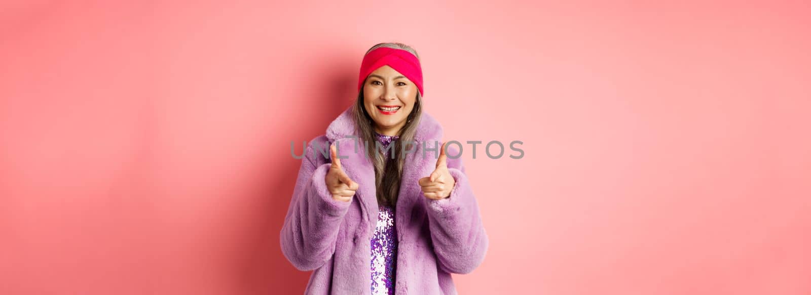 Fashion and shopping concept. Cool asian senior woman in stylish fake fur coat pointing fingers at camera, asking you to check out promo offer, standing over pink background.