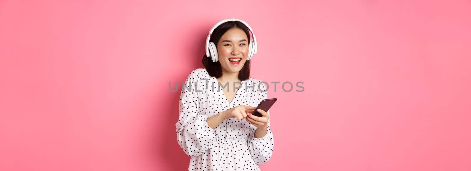 Beautiful asian woman listening music in headphones, using mobile phone, smiling happy at camera, standing over pink background by Benzoix