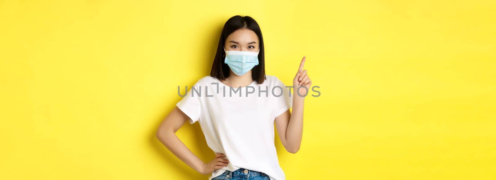 Covid, health care and pandemic concept. Asian female model in medical mask and white t-shirt pointing finger at upper left corner logo, showing promotion, yellow background.