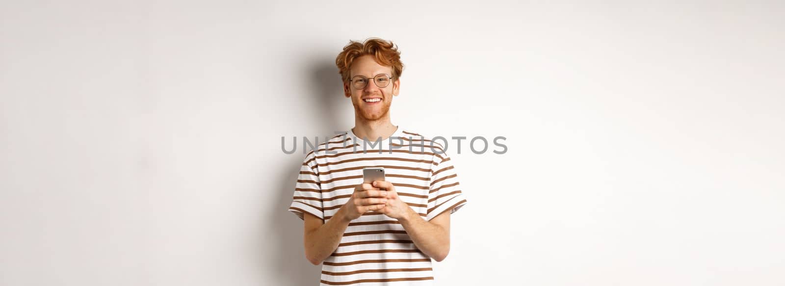 Technology and e-commerce concept. Redhead guy in glasses using mobile phone and smiling. Young man with smartphone staring happy at camera, white background.