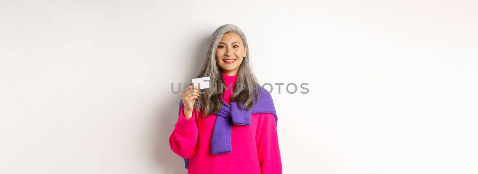 Shopping concept. Smiling asian middle-aged woman with grey hair showing plastic credit card and looking happy, standing over white background.