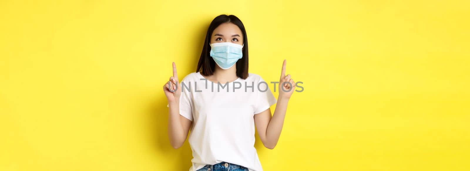 Covid-19, pandemic and social distancing concept. Young asian woman in white t-shirt and medical mask from coronavirus, looking and pointing fingers up, showing special offer.