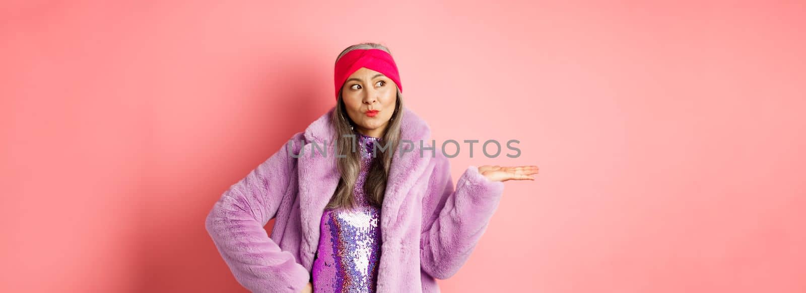 Fashion and shopping concept. Stylish asian senior woman in purple winter outfit demonstrating product on hand. Cool old lady in trendy clothes holding something on palm, pink background by Benzoix
