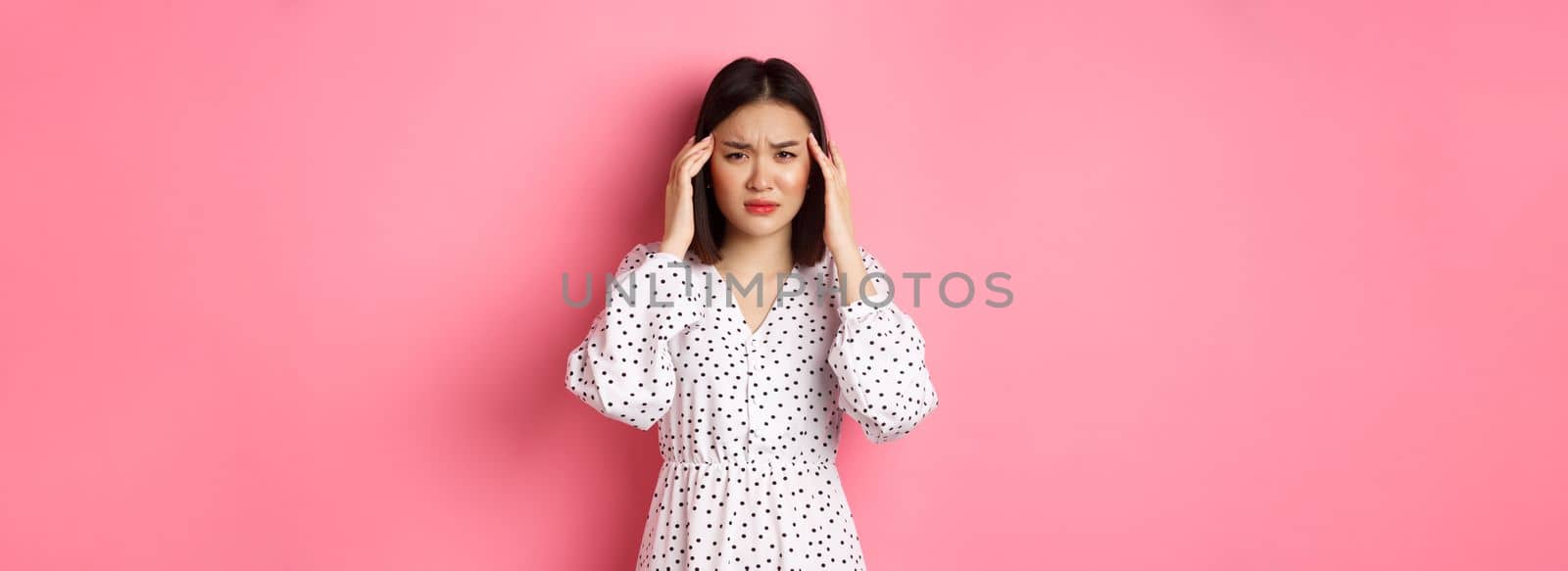 Image of upset asian woman having headache, feeling unwell or dizzy, close eyes and massaging head, suffering migraine, standing over pink background.