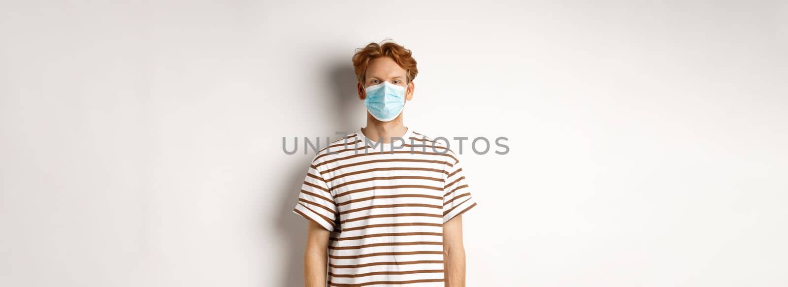 Covid-19, pandemic and social distancing concept. Young man with red hair wearing medical mask to prevent catching coronavirus, white background.