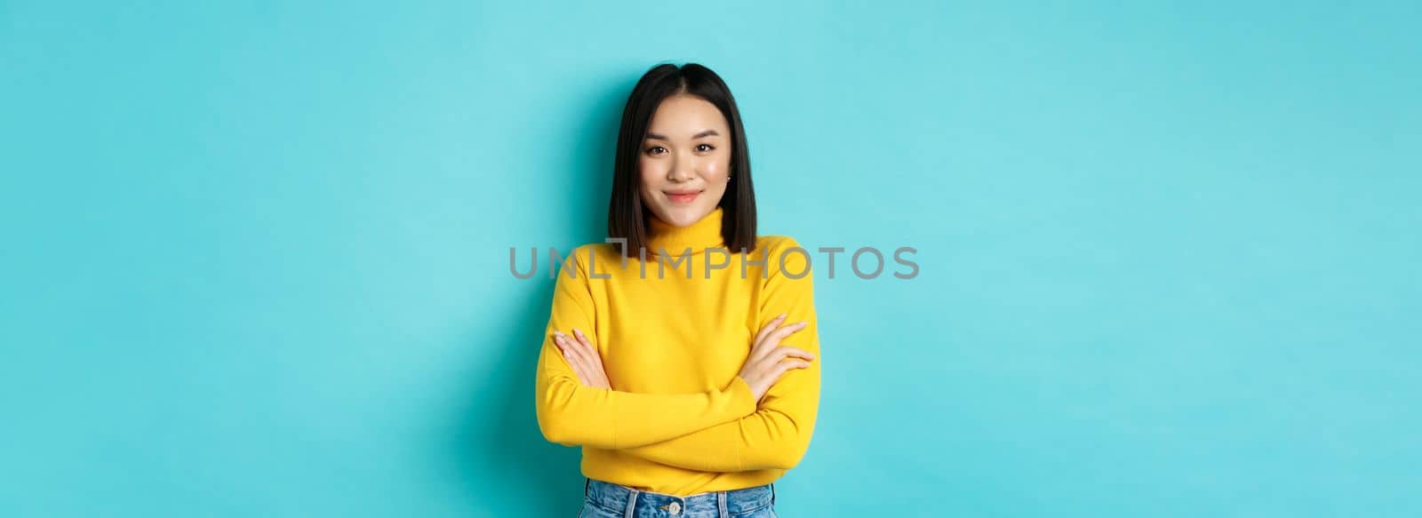 Attractive asian woman with short dark hair, cross arms on chest and smiling confident, standing over blue background.