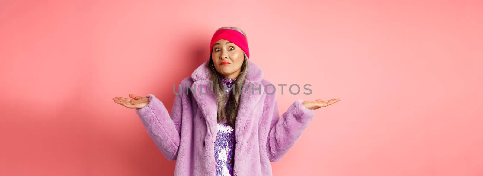 Fashionable and funky asian old woman shrugging shoulders, looking confused and clueless, standing in stylish purple coat and headband, pink background.