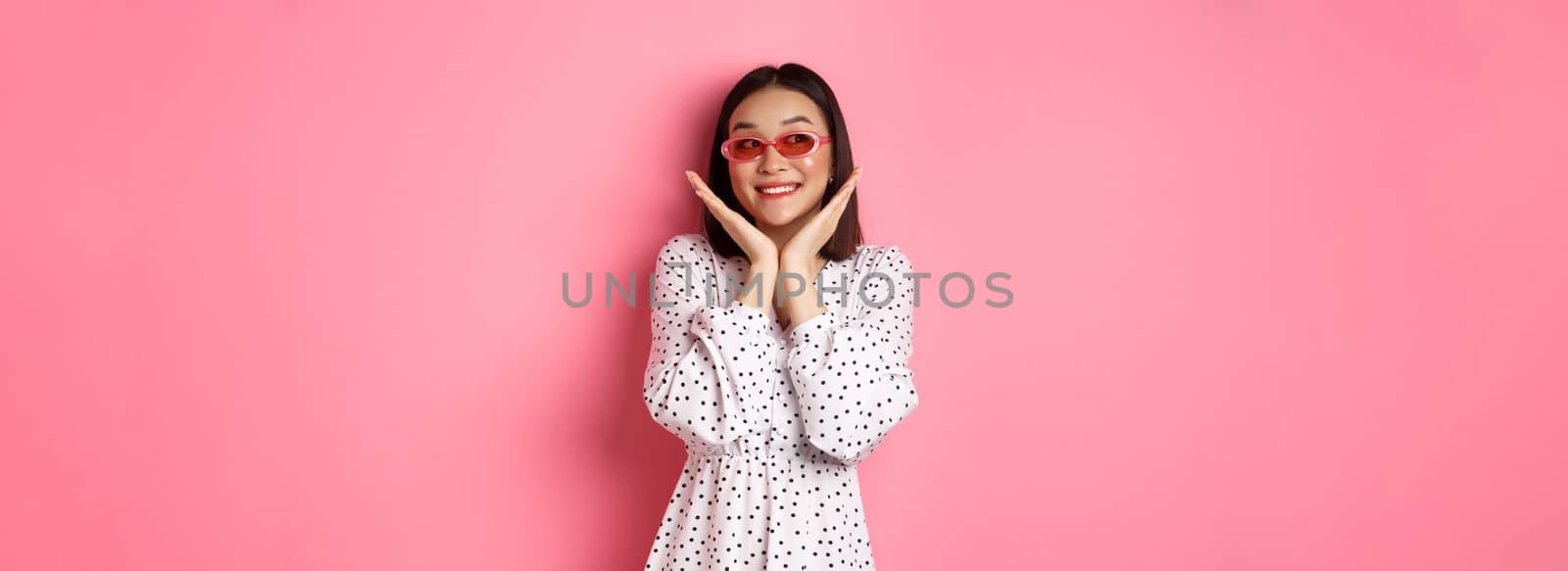 Tourism and lifestyle concept. Beautiful asian woman showing her clean cute face, wearing sunglasses, smiling and looking aside with admiration, standing over pink background by Benzoix