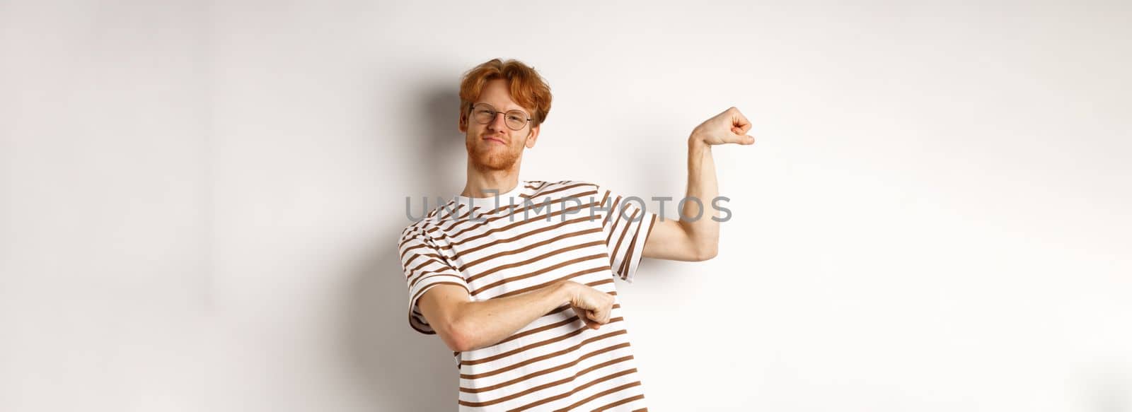 Image of confident and strong redhead man flexing biceps, showing muscles after gym, standing over white background by Benzoix