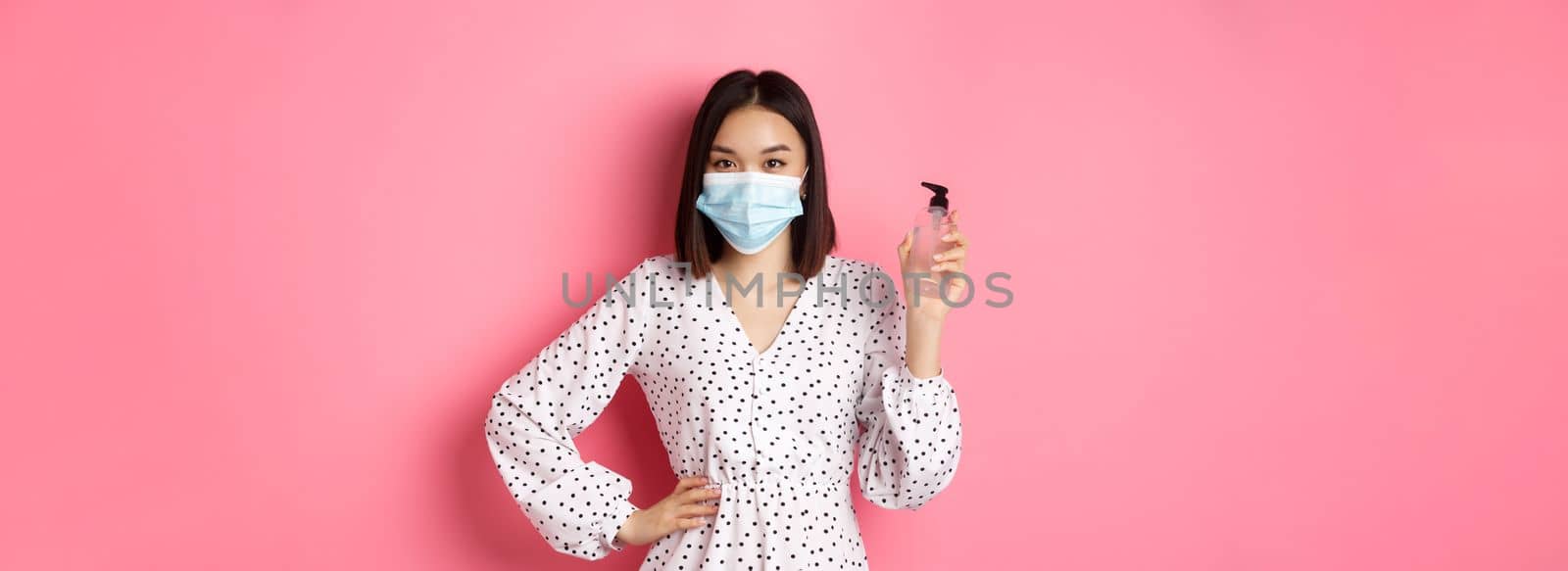 Covid-19, pandemic and lifestyle concept. Beautiful korean woman in dress and medical mask showing hand sanitizer, recommending antiseptic, standing over pink background by Benzoix