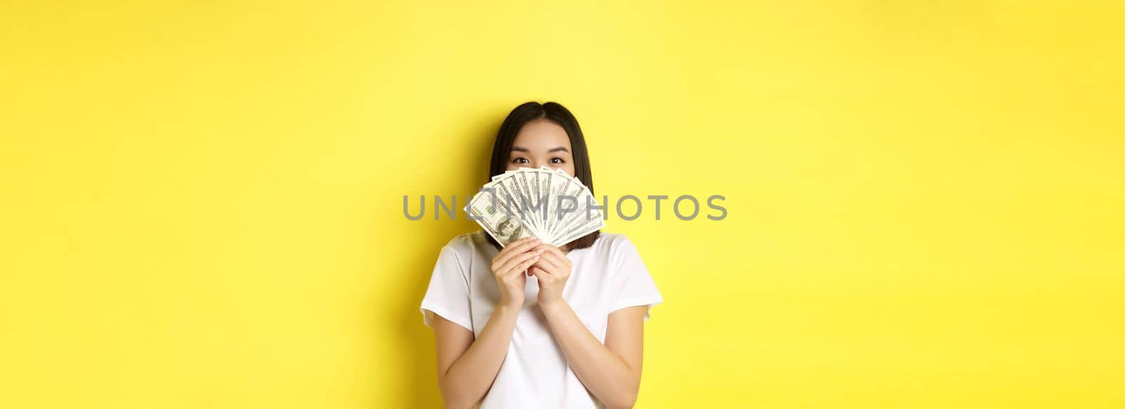 Cute asian woman hiding face behind money, peeking at camera satisfied, earn cash, standing over yellow background.