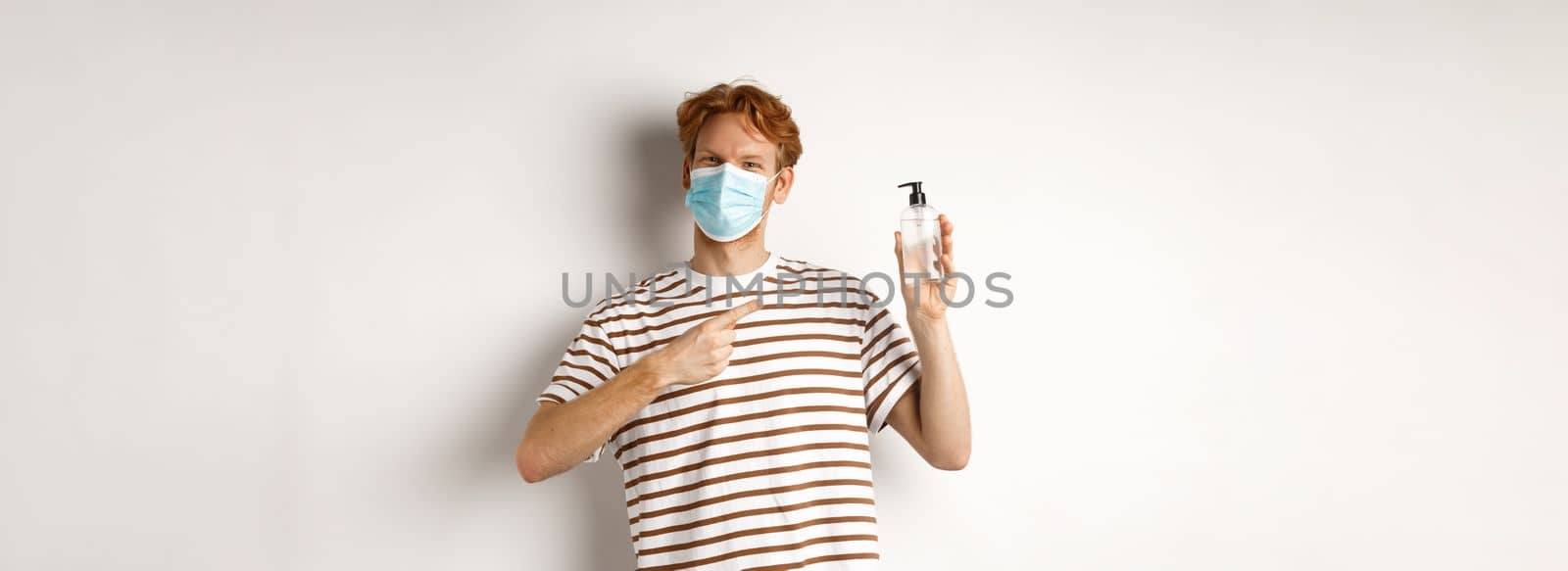Covid-19, health and lifestyle concept. Cheerful redhead man in face mask pointing finger at hand sanitizer, recommending antiseptic, white background.