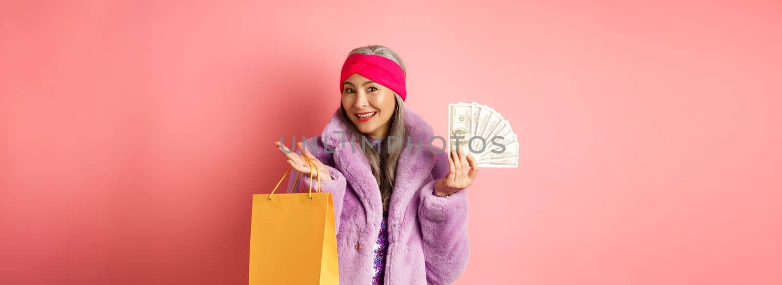 Rich and stylish grandmother going on shopping with dollars, holding paper bag and smiling carefree, standing over pink backgrund.