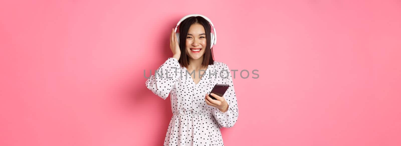 Beautiful asian woman listening music in headphones, using smartphone streaming app and smiling, standing over pink background.