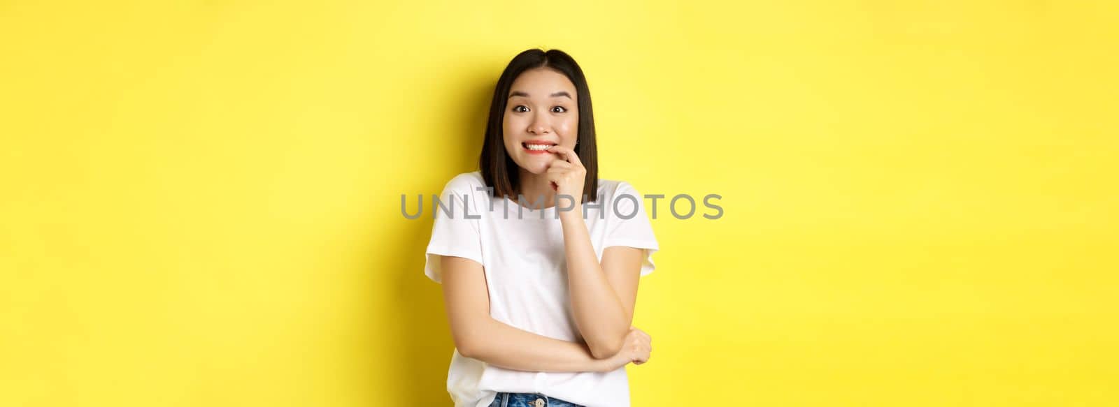 Portrait of cheerful asian girl wants something, looking with desire and temptation at camera, smiling at camera, standing ovr yellow background by Benzoix