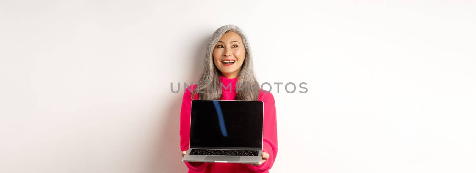 E-commerce concept. Happy beautiful asian senior woman with grey hair, looking aside and smiling, showing laptop black screen, standing over white background.