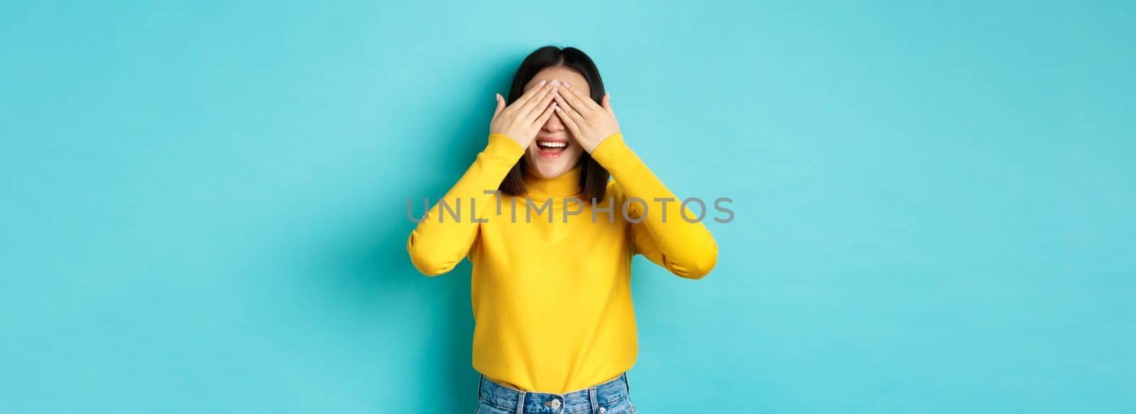 Cheerful asian girl in yellow pullover waiting for surprise, playing hide n seek and smiling, expecting gift with eyes closed, standing over blue background by Benzoix