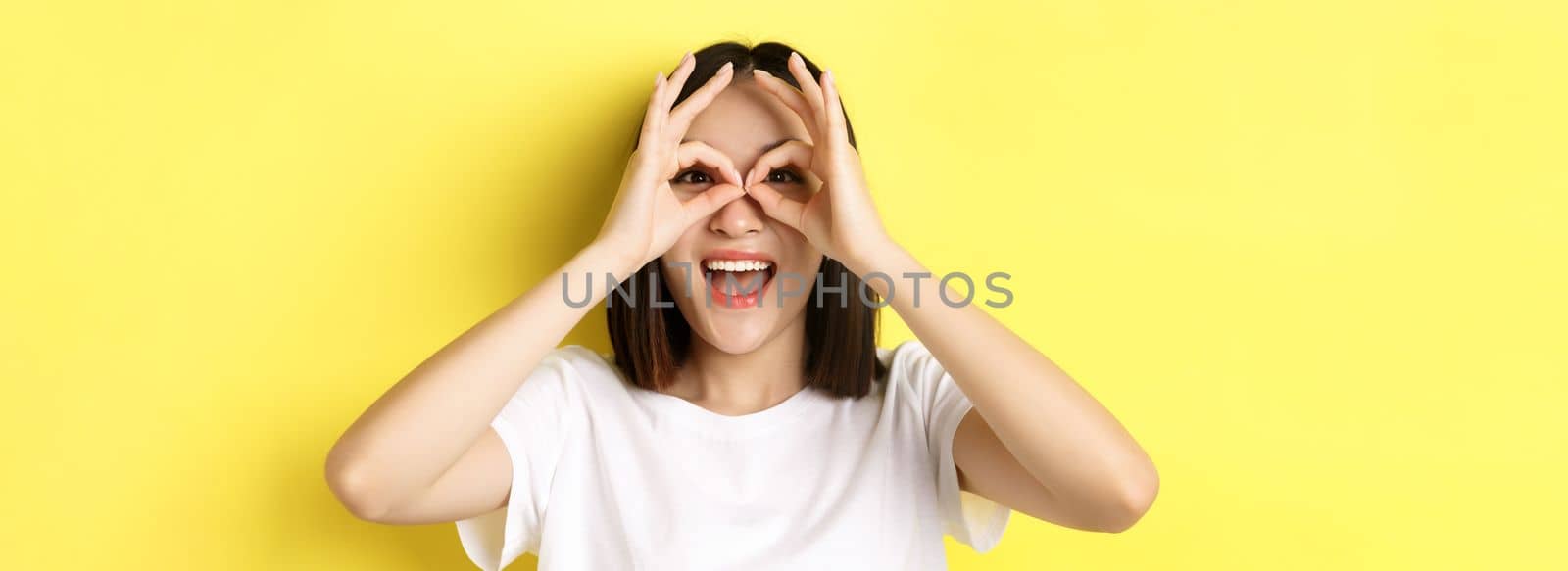 Close up of funny asian girl looking through hand binoculars and smiling, standing in t-shirt against yellow background by Benzoix