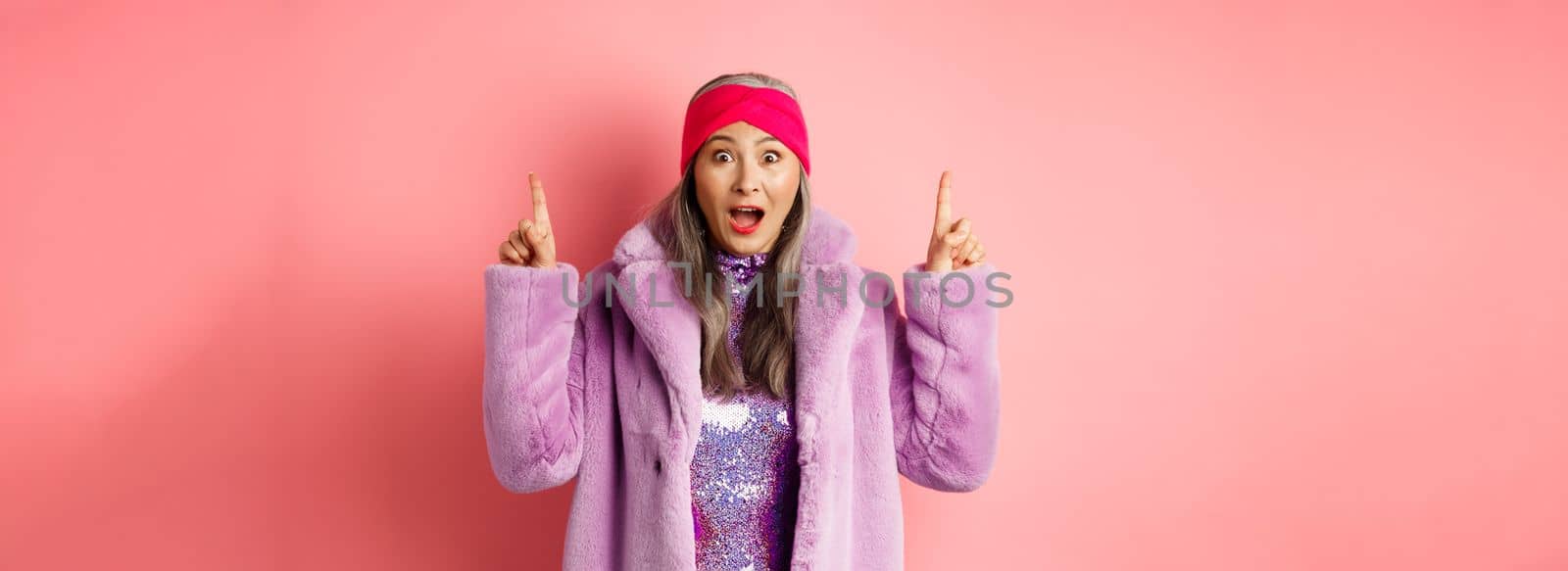 Happy elderly asian lady pointing fingers up, looking amazed at camera, showing cool promo offer, standing against pink background in purple fake fur coat.