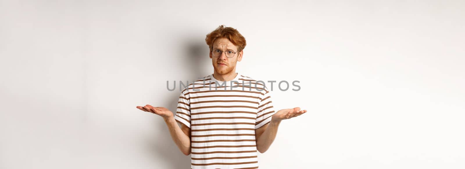 Confused boyfriend with red hair shrugging, spread hands sideways and staring at camera puzzled, asking what, stnading over white background.