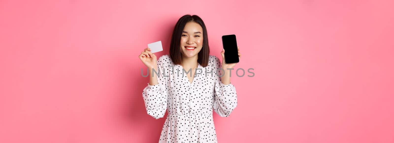 Joyful korean woman showing smartphone screen and credit card, paying for internet order, demonstrating online shopping app, standing over pink background by Benzoix