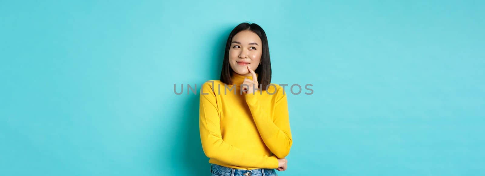 Beautiful korean girl thinking, imaging and smiling, looking at upper left corner and daydreaming, standing against blue background.