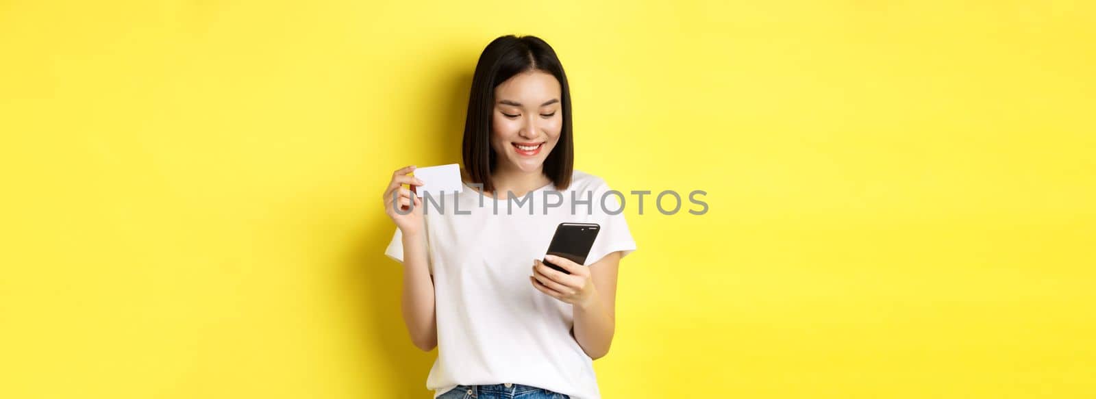 E-commerce and online shopping concept. Beautiful asian woman paying in internet, looking at smartphone screen and holding plastic credit card, yellow background.