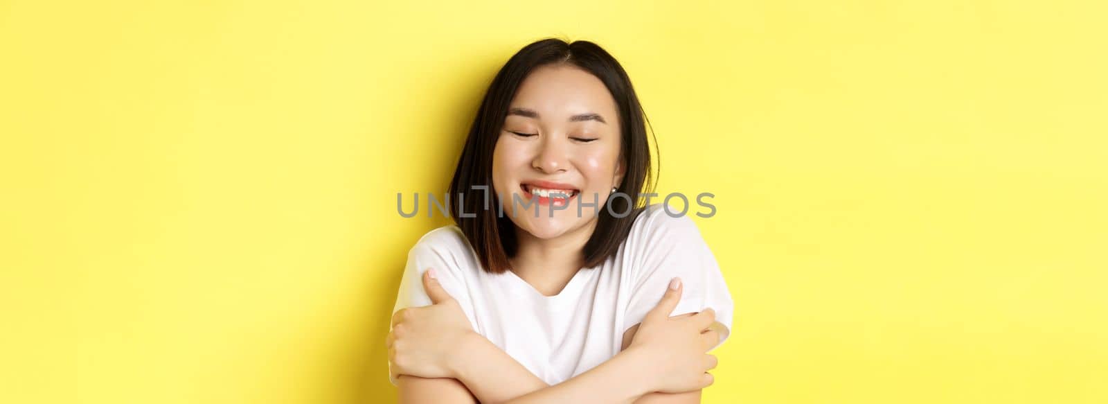 Close up of romantic asian girl hugging herself and dreaming, close eyes and smile while imaging something tender, standing over yellow background by Benzoix