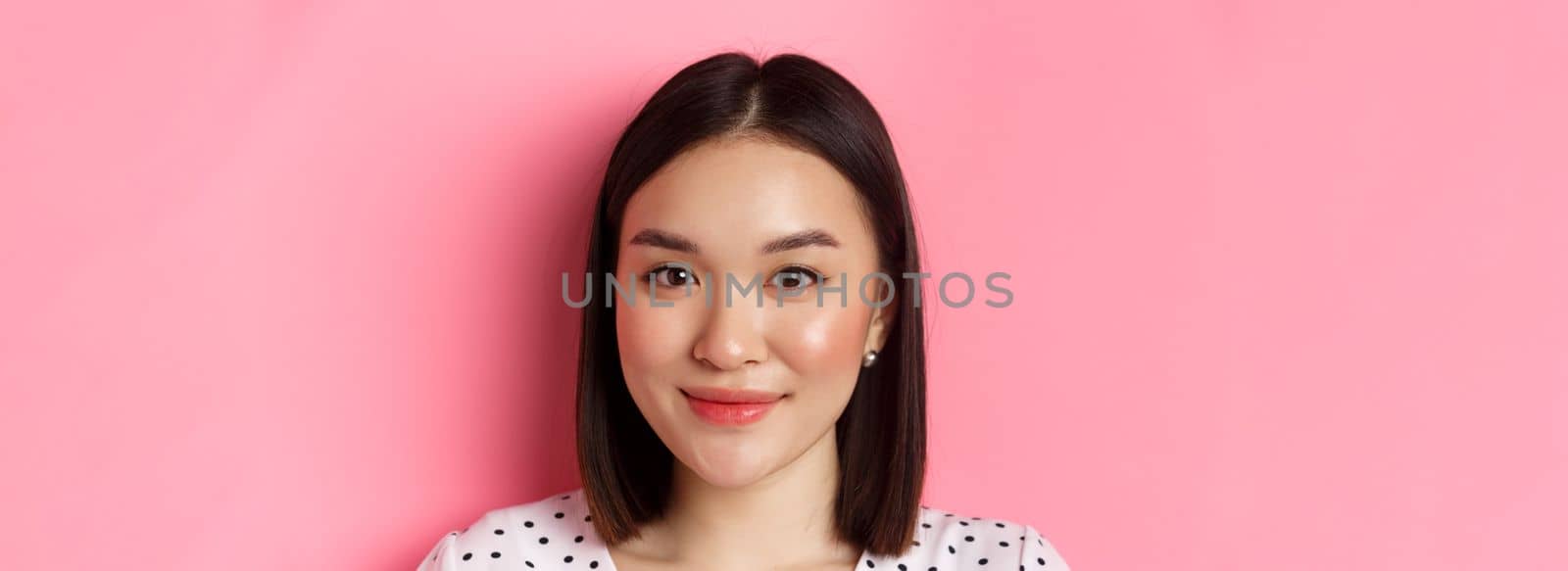 Beauty and lifestyle concept. Headshot of beautiful asian woman smiling, looking at camera happy and romantic, standing against pink background.