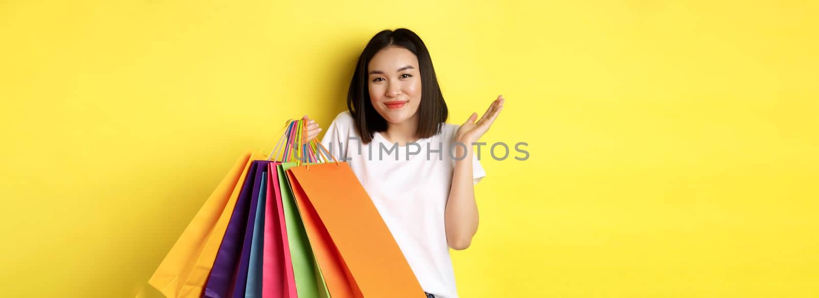Silly asian girl showing shopping bags and shrugging, shop in stores with discounts, standing over yellow background by Benzoix