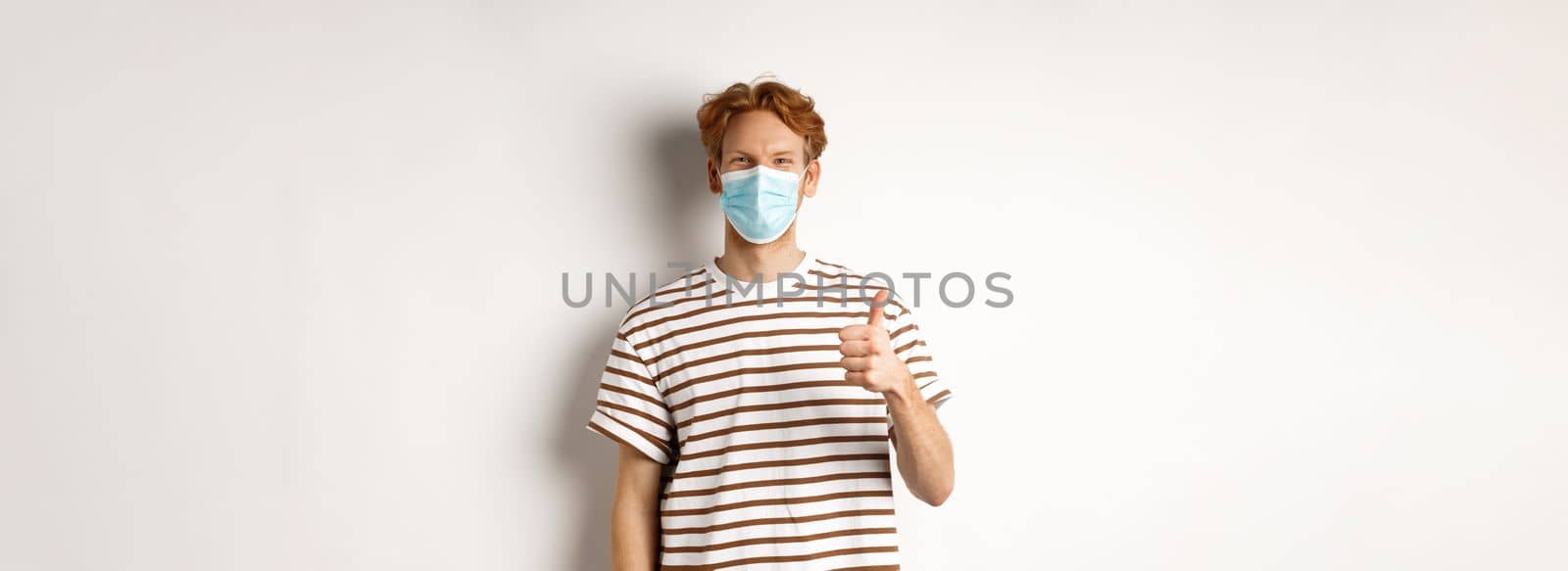 Covid-19, pandemic and social distancing concept. Young man with red hair wearing medical mask to prevent catching coronavirus, showing thumbs-up, white background.