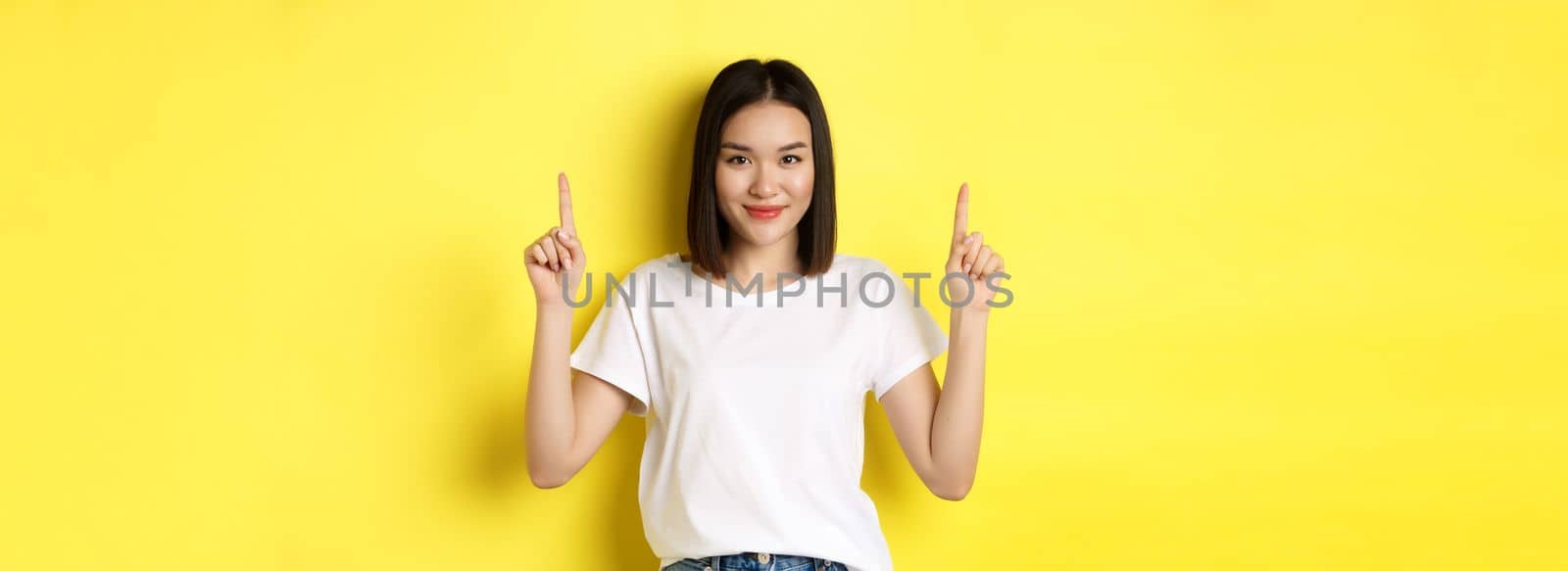 Beauty and fashion concept. Beautiful asian woman in white t-shirt pointing fingers up, standing over yellow background.