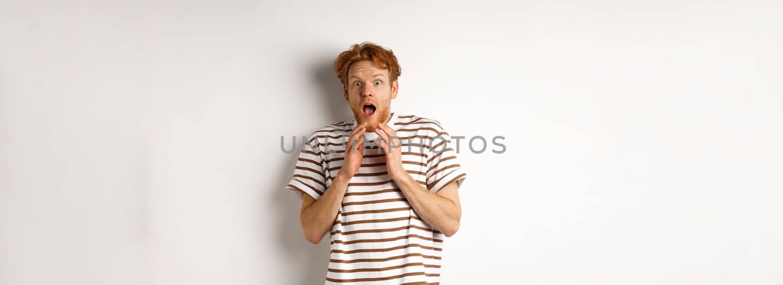 Timid young man with red hair, looking scared, jumping and screaming from fear, standing over white background.