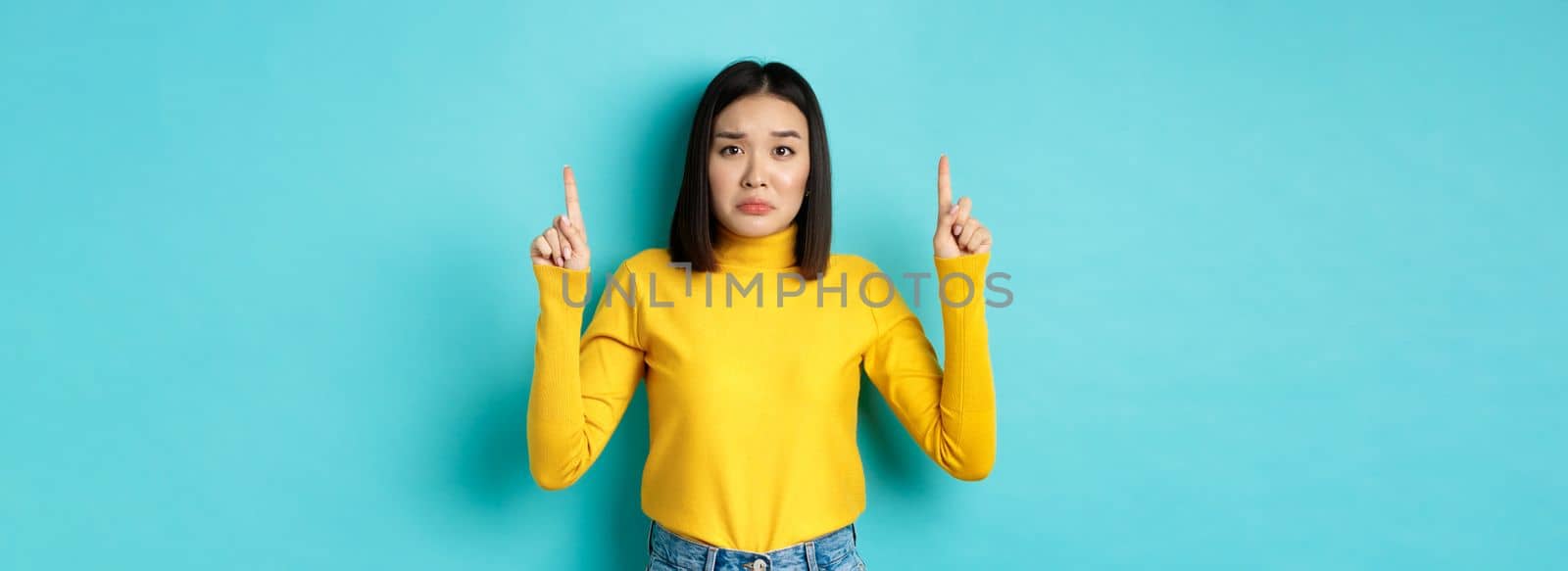 Shopping concept. Disappointed and gloomy asian woman sulking upset, pointing fingers up at bad news banner, standing over blue background by Benzoix